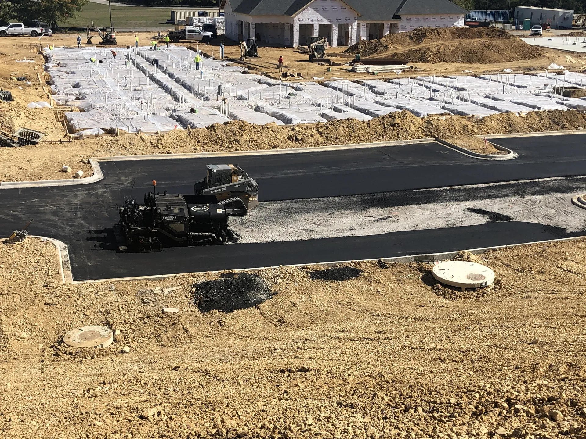 Aerial view of a Project One Contracting paver and skid steer laying a new asphalt road through a residential development under construction