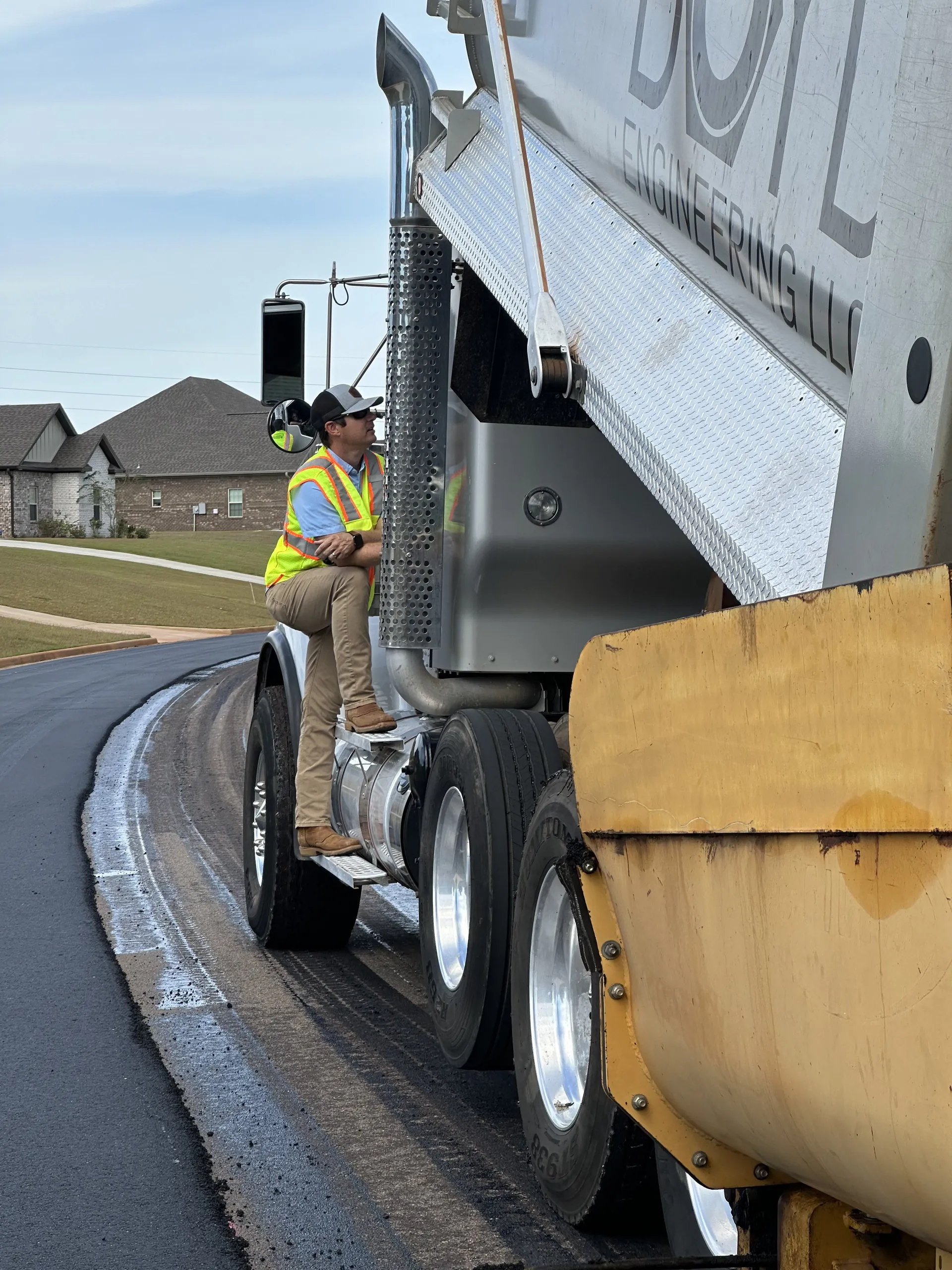 Graham Wiseman, Project One Contracting operations lead, riding the step of a dump truck while directing a fresh asphalt paving job on a curved residential street