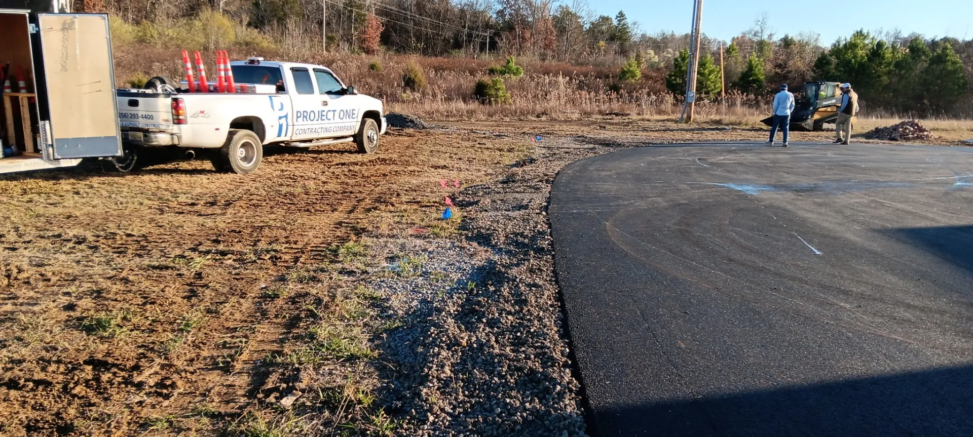 Project One Contracting pickup truck and trailer parked beside a newly paved asphalt curve with crew working in the background