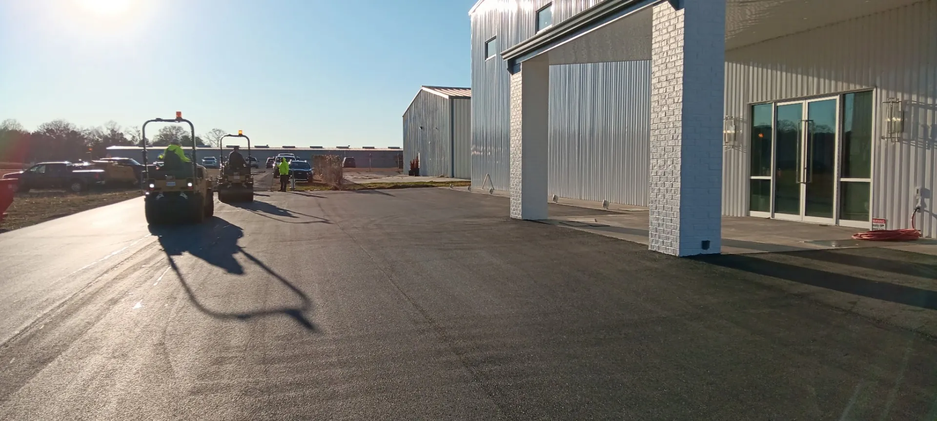 Two Project One Contracting roller operators compacting fresh asphalt along a commercial building drive on a sunny morning